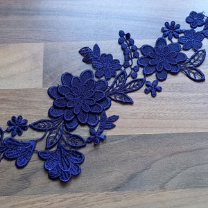 May include: A navy blue floral lace applique with detailed flower and leaf designs. The applique is made of delicate, openwork lace, ideal for embellishing garments or craft projects. The intricate details are visible against a wooden background.