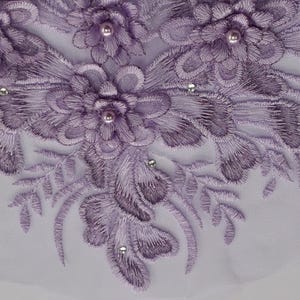 May include: Close-up of lavender floral lace fabric. The design features embroidered flowers with pearl centres and sparkling rhinestone accents. The lace is set against a white background, creating a delicate and elegant look.