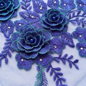 May include: Close-up of a floral lace design featuring embroidered blue roses with teal accents and small, sparkling embellishments. The design includes leaves and smaller flowers, all on a light blue background.