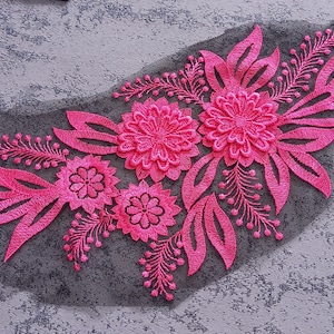 May include: A bright pink floral embroidered applique on a dark grey mesh base. The design features several flowers and leaves, suitable for embellishing clothing or craft projects. The applique is a decorative element.