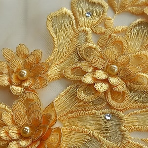 Pode incluir: Close-up de renda floral dourada bordada. O design apresenta flores detalhadas com centros perolados e pequenos enfeites brilhantes. A renda é provavelmente usada para embelezar roupas ou artesanato.