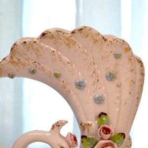 May include: A vintage, fan-shaped ceramic vase in a soft pink hue, adorned with gold accents and delicate blue floral details. The vase features a curved handle and is embellished with small pink roses and green leaves.