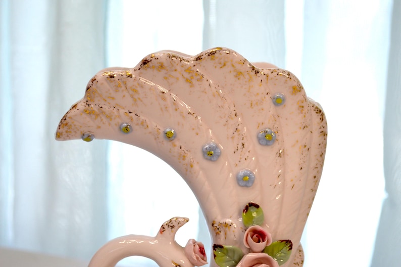 May include: A vintage, fan-shaped ceramic vase in a soft pink hue, adorned with gold accents and delicate blue floral details. The vase features a curved handle and is embellished with small pink roses and green leaves.