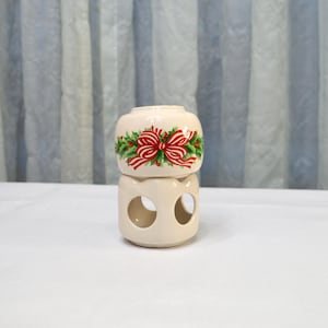 May include: A cream-colored ceramic candle holder with a festive design. The top section features a red bow with green holly accents. The bottom section has two circular cutouts. The candle holder is on a white surface.