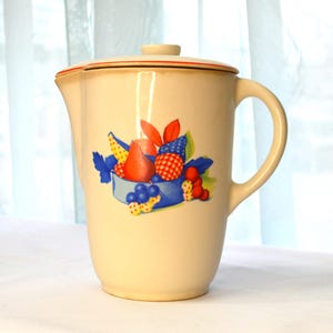 May include: A white ceramic jug with a red rim and lid. The jug features a fruit design with pears, grapes, and strawberries in shades of red, blue, and yellow.