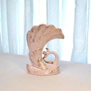 May include: A vintage ceramic swan vase in a soft pink hue, adorned with gold accents. The swan features a curved neck and a stylized tail, with delicate floral details. The vase is a decorative item.