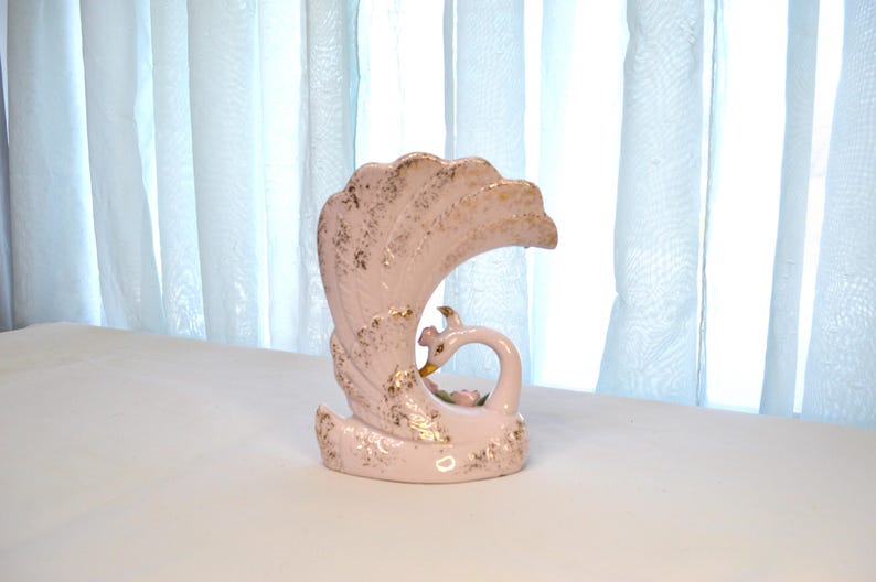 May include: A vintage ceramic swan vase in a soft pink hue, adorned with gold accents. The swan features a curved neck and a stylized tail, with delicate floral details. The vase is a decorative item.