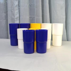 May include: A collection of tall, stackable plastic tumblers in various colors: navy blue, white, and yellow. The tumblers have a two-part design with a slightly flared top and a ridged base. The background is a light-colored surface and a white curtain.