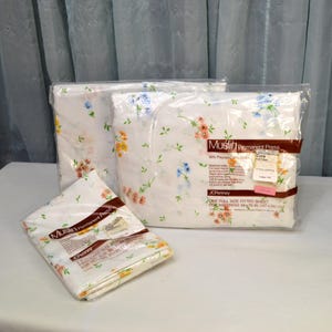May include: Three packages of JCPenney Muslin Permanent Press sheets. The sheets are white with a floral pattern in shades of orange, blue, and yellow. The packages are sealed in clear plastic and are on a white surface.