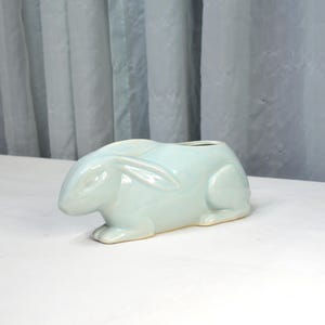 May include: A light blue ceramic bunny-shaped planter. The planter has a rectangular opening on top and is designed in the form of a reclining rabbit. The bunny has detailed ears and paws. The planter is on a white surface.