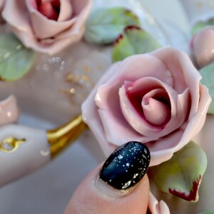 May include: Close-up of a white ceramic decorative piece adorned with pink roses, green leaves, and gold accents. The roses have a detailed, realistic appearance. A hand with a black and white speckled nail is in the foreground.