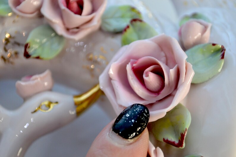 May include: Close-up of a white ceramic decorative piece adorned with pink roses, green leaves, and gold accents. The roses have a detailed, realistic appearance. A hand with a black and white speckled nail is in the foreground.