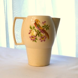 May include: A white ceramic pitcher with a bird and floral design. The pitcher has a wide mouth and a handle.