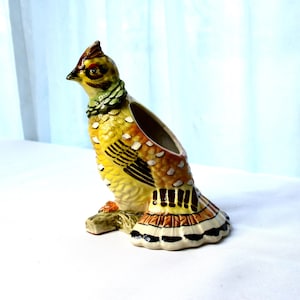 May include: A ceramic bird figurine with a yellow, brown, and green painted design. The bird is perched on a branch and has a small opening in its back, suggesting it may be a planter or a decorative container.