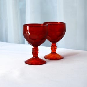 May include: Two red glass goblets with a faceted design. The goblets have a short stem and a wide bowl.