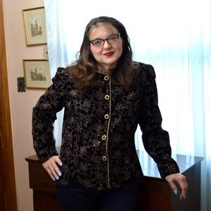 May include: Black velvet jacket with gold floral embroidery and gold trim. The jacket has a round collar and is fastened with gold buttons. The sleeves are long and slightly puffed at the shoulders. The jacket is worn over dark blue pants.