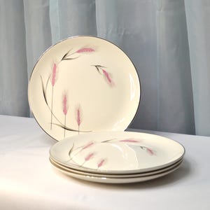 May include: Three white ceramic dinner plates with a pink and black wheat design. The plates have a gold rim.