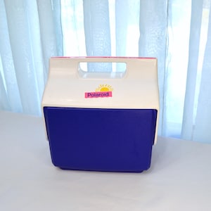 May include: A vintage white and blue cooler with a handle. The cooler has a blue base and a white top with a pink Polaroid sticker featuring a sun graphic. The cooler is designed to keep items cold.