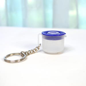 May include: A miniature clear plastic measuring cup with a blue lid and handle, attached to a silver keychain. The cup has printed measurement markings. The keychain is made of a chain and a split ring.