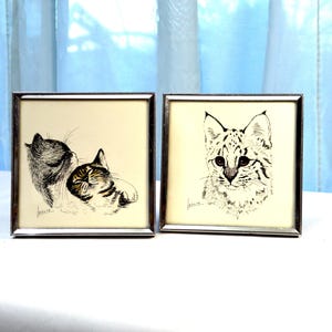 May include: Two framed cat art prints. One print features two cats, one grooming the other. The second print is a close-up of a cat's face with spotted markings. Both prints are in silver-colored frames.
