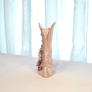 May include: A tall, light pink ceramic vase with a textured surface and a unique shape. The vase is adorned with delicate pink roses and gold accents. The vase is approximately 10 inches tall.