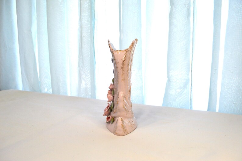 May include: A tall, light pink ceramic vase with a textured surface and a unique shape. The vase is adorned with delicate pink roses and gold accents. The vase is approximately 10 inches tall.
