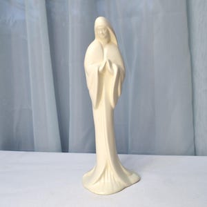 May include: White ceramic figurine of a woman in a long robe with her hands clasped in prayer.