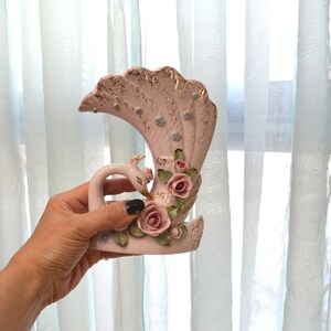 May include: A vintage ceramic vase in the shape of a swan with a fan-shaped back. The vase is primarily pink with gold accents and features pink roses and green leaves. The swan has a long neck and a detailed head.