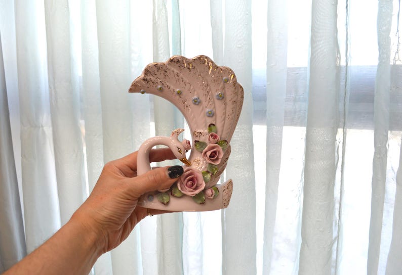 May include: A vintage ceramic vase in the shape of a swan with a fan-shaped back. The vase is primarily pink with gold accents and features pink roses and green leaves. The swan has a long neck and a detailed head.