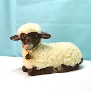 May include: A figurine of a lamb in a resting position, featuring a dark brown face and legs with a textured, cream-colored faux fur body. The lamb wears a thin gold-colored cord around its neck with a small, metallic bell attached at the front.