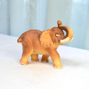May include: A brown ceramic elephant figurine with its trunk raised in the air. The elephant has a light brown body and a darker brown trunk.