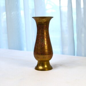May include: A small, brass vase with an ornate design. The vase is a light gold colour and has a flared top and a round base.