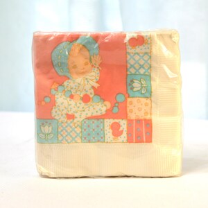 以下が含まれることがあります： ベビーのデザインが施された、パステルピンクとブルーの紙ナプキンのパッケージ。デザインは、青いボンネットをかぶってビーズの紐を持っている赤ちゃん。背景はパッチワークのキルトデザインです。