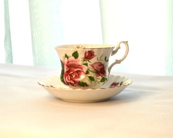 May include: A white teacup and saucer with a floral pattern of pink roses and green leaves. The teacup has a delicate handle and a scalloped rim.