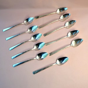 May include: Ten silver teaspoons with ornate handles arranged in a fan shape on a light pink background.