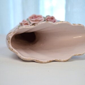 May include: A ceramic vase in a horn shape, with a light pink interior and exterior. The rim has a scalloped edge with gold trim. Pink roses with green leaves are at the top of the vase.