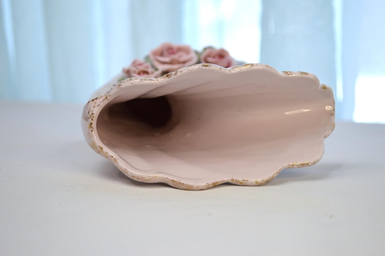 May include: A ceramic vase in a horn shape, with a light pink interior and exterior. The rim has a scalloped edge with gold trim. Pink roses with green leaves are at the top of the vase.