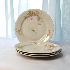May include: Set of four white porcelain plates with a delicate floral pattern in shades of pink and green. The plates have a scalloped edge and are perfect for a vintage or romantic table setting.