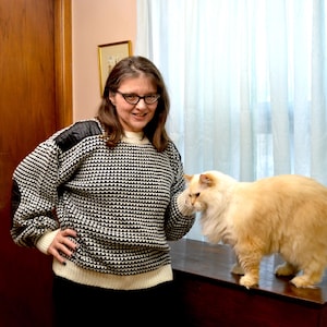 May include: A woman wearing a black and white patterned sweater with black accents on the shoulders and elbows. A fluffy, light orange cat stands on a dark brown surface next to the woman.