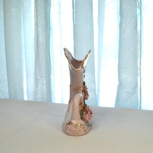 May include: A decorative, light pink ceramic vase with a handle and floral accents. The vase features a flared top and is adorned with pink roses and gold detailing. The vase is approximately 8 inches tall.