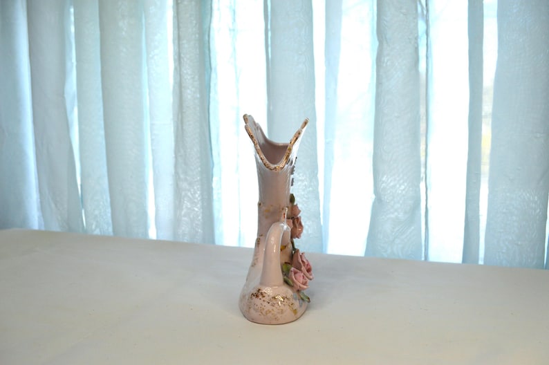 May include: A decorative, light pink ceramic vase with a handle and floral accents. The vase features a flared top and is adorned with pink roses and gold detailing. The vase is approximately 8 inches tall.