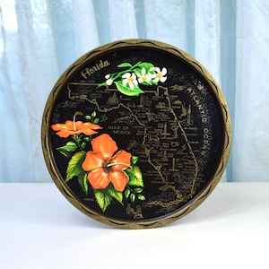 May include: Circular black tray with a gold-coloured rim, depicting a map of Florida. The tray is adorned with orange hibiscus flowers, white flowers, and the word "Florida" in gold lettering. The Gulf of Mexico and Atlantic Ocean are labelled.
