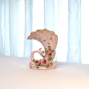 May include: A ceramic peacock vase in a soft pink hue, adorned with delicate gold accents and clusters of pink roses. The vase features a swan-like form with a fanned tail, creating an elegant and decorative piece.