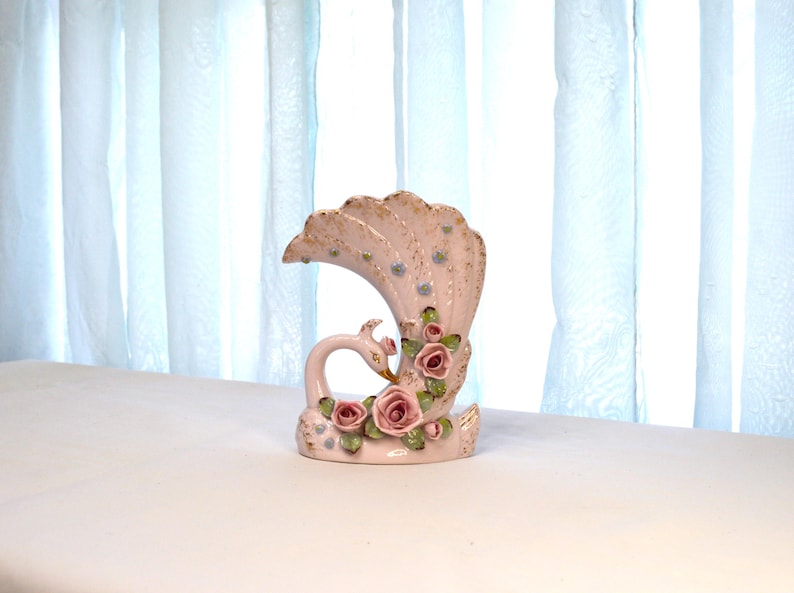 May include: A ceramic peacock vase in a soft pink hue, adorned with delicate gold accents and clusters of pink roses. The vase features a swan-like form with a fanned tail, creating an elegant and decorative piece.
