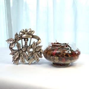 May include: A silver metal Christmas ornament featuring a lantern with a bow and poinsettia leaves. A glass bowl filled with potpourri sits next to the ornament.