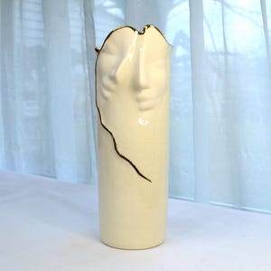 May include: A white ceramic vase with a gold trim and a face design. The vase is tall and slender.
