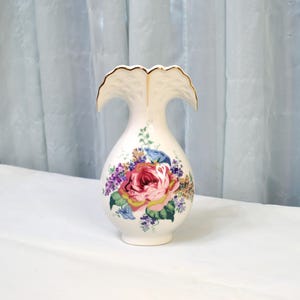 May include: A cream-colored ceramic vase with a ruffled top and gold trim. The vase features a large, colorful floral design with a prominent pink rose, surrounded by other flowers and green leaves. The vase is on a white surface.