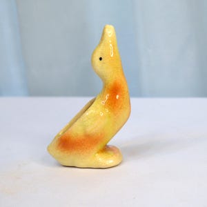 Puede incluir: Una figurita de pato de cerámica con un esmalte amarillo y naranja. El pato está sentado con la cabeza echada hacia atrás.