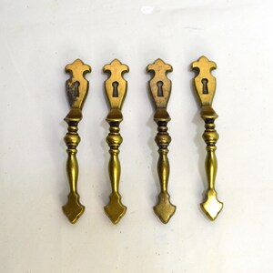 May include: Four matching brass drawer pulls with a vertical orientation. Each pull features a decorative top plate with a keyhole cutout, a slender central stem with rounded detailing, and a pointed bottom finial. The metal has a polished finish.