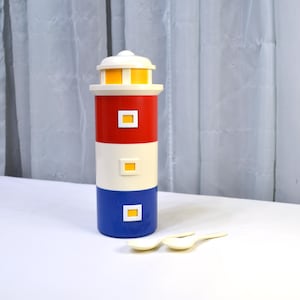 May include: A colorful lighthouse-shaped container with a white top and base, a red middle section, and a blue bottom. The container has small yellow square windows. Two small, off-white spoons are in front of the container.